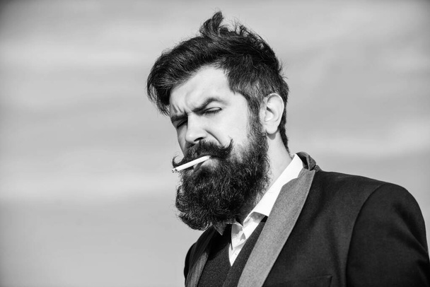 Truth about smoking pleasure and nicotine addiction. Bearded hipster smoking cigarette blue sky background. Guy with cigarette enjoy nicotine influence. Man with beard and mustache hold cigarette - 写真・画像
