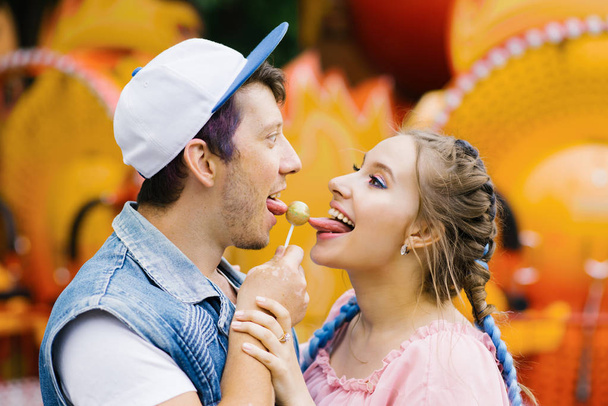 A cheerful happy couple of a guy and a girl eat candy on a stick, smile and stick out their tongues to a Lollipop in an amusement Park - Photo, Image