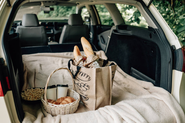 Picknickpaket und Korb mit Croissant und Kaffee und Popcorn liegen auf dem Plaid im Kofferraum des Autos - Foto, Bild