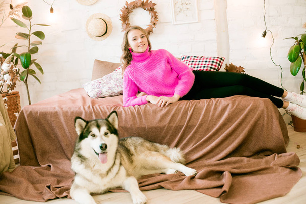 Cute teenage girl with blond curly hair at home, on the bed in the loft with a big malamute dog. People and Dogs. - Photo, image