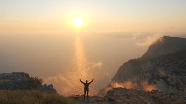 Silhouette of a Young Woman standing on a cliff edge observing a beautiful dramatic sunset over a sea from a high mountain in Crimea. Lady hiker with arms outstretched against beautiful sunset light. - Video, Çekim