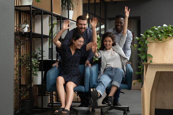 Overjoyed young diverse colleagues having fun, riding on chairs. - Foto, Imagem