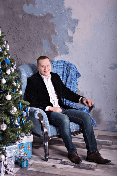 elegante man van middelbare leeftijd zit in de buurt van een nieuwjaarsboom in een pak voor Kerstmis - Foto, afbeelding