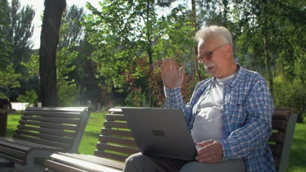 Naštvaný starší muž mluví skypem, sedí na lavičce v zahradě. Důchodce má na klíně laptop. Mává rukou, někomu nadává - Záběry, video
