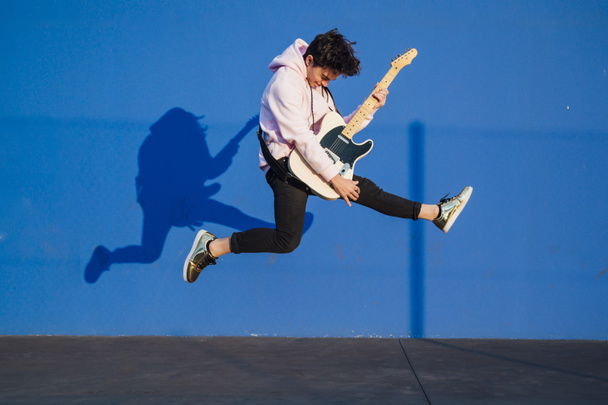 Junger Mann springt mit E-Gitarre auf blauem Hintergrund - Foto, Bild