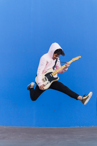 Junger Mann springt mit E-Gitarre auf blauem Hintergrund - Foto, Bild
