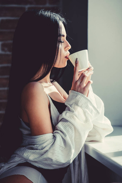 The best coffee for her daily routine. Sensual girl drinking her favorite morning coffee. Pretty woman drinking fresh hot coffee at window. Sexy girl holding ceramic coffee cup - Photo, Image