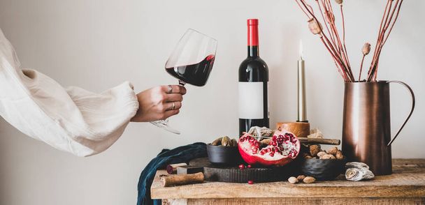 Red wine, board with cheese, fruit, almonds and olives, candles, decorative flowers on kitchen counter and womans hand holding glass of wine, white wall at background, wide composition - Foto, Imagem