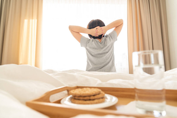 Tálca vízzel és keksszel reggeli az ágyon, nő nyújtózkodik a háttérben - Fotó, kép