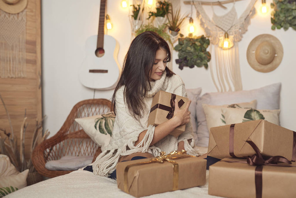 christmas, happy boho girl wraps presents on the bed, bright and festive atmosphere, brunette - Фото, изображение