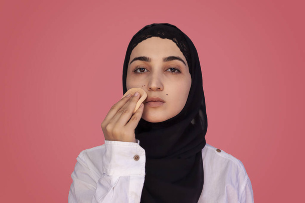 Leuke twintiger jaren vrouw die poeder met spons op haar gezicht aanbrengt. Mooie Perzische vrouw in hijab doet make-up - Foto, afbeelding