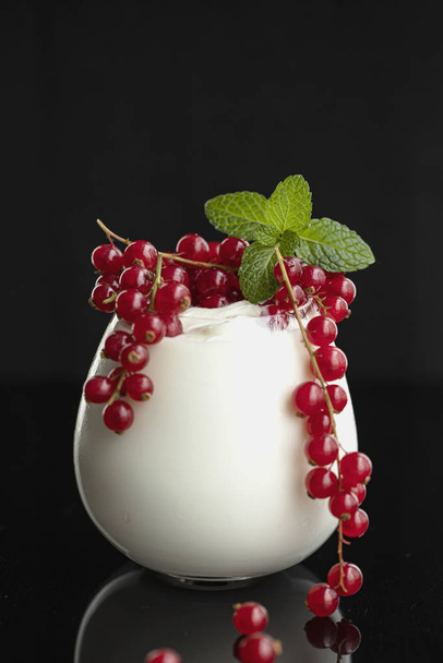 Natural yogurt in a glass is decorated with fresh red currant berries and mint leaves on black background. - Foto, Bild