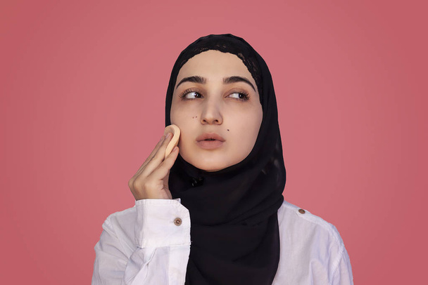 Linda hembra de 20 años aplicando polvo con esponja en su cara. Hermosa mujer persa en hiyab haciendo maquillaje
 - Foto, Imagen