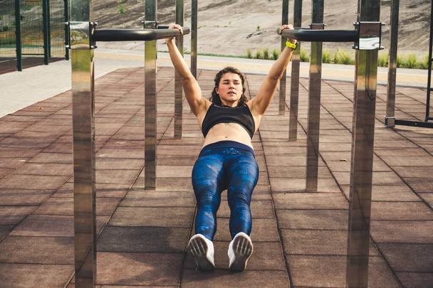 girl trains outdoors in street gym. Training of biceps and triceps. woman parallel bars workout exercise. female athlete exercising on parallel bars, doing push-ups. Sport and bodybuilding concept - Photo, Image
