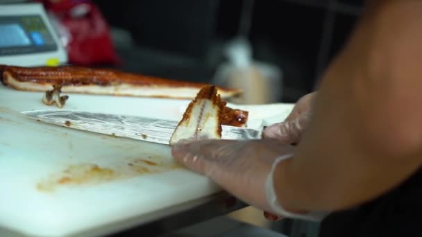 Hombre asiático en guantes estériles transparentes hábilmente cortar sabroso pescado frito con un cuchillo afilado para la venta en una mesa blanca en la cocina de cerca. Maestro del sushi cocinando deliciosos rollos
. - Metraje, vídeo