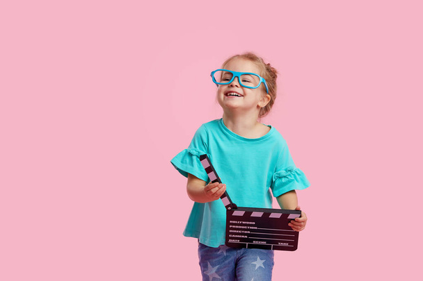 Lustig lächelndes Kindermädchen mit Kinobrille hält Filmklapptafel isoliert auf rosa Hintergrund. Studioporträt. Lifestyle-Konzept für Kinder. Kopierraum für tex - Foto, Bild