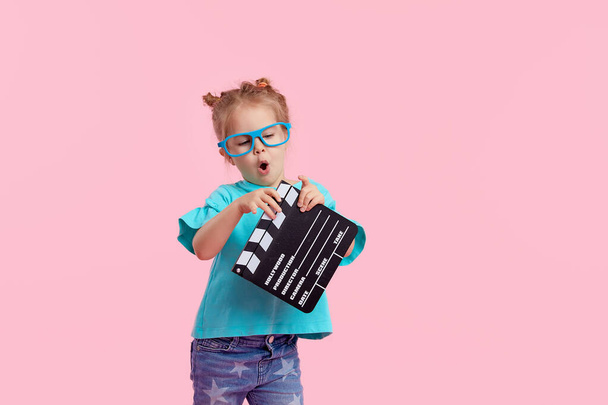 Funny smiling child girl in cinema glasses hold film making clapperboard isolated on pink background. Studio portrait. Childhood lifestyle concept. Copy space for tex - Foto, afbeelding