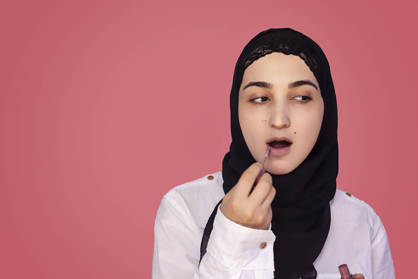 Lindo 20s hembra poner lápiz labial con un cepillo. Hermosa mujer persa en hijab haciendo maquillaje aplicando lápiz labial en sus labios
 - Foto, Imagen