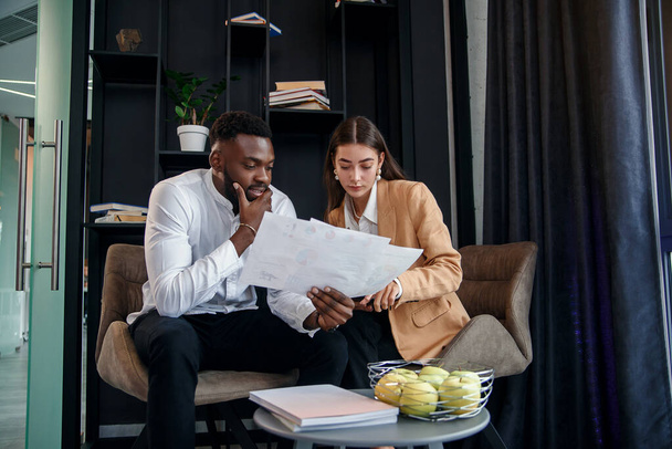 Business couple of african american man and european woman which sitting in specially designated room and using paper documents and tablet pc, conversing about their business project and their - Fotoğraf, Görsel