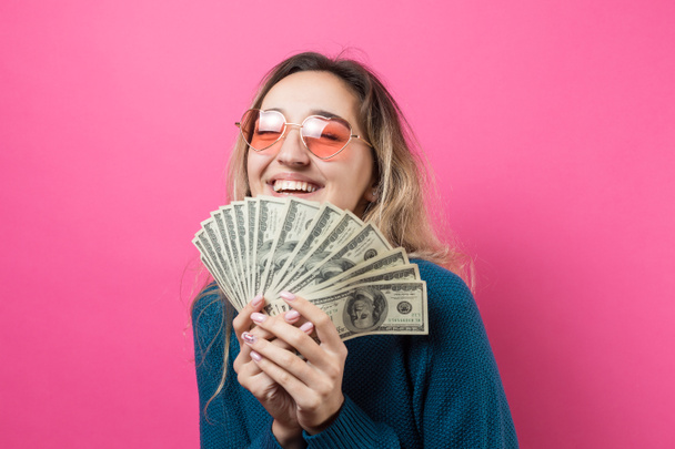 Κοντινό πλάνο της νεαρής όμορφης γυναίκας με ένα μπλε πουλόβερ σε γυαλιά με αμερικανικά δολάρια στο χέρι πάνω από ροζ φόντο. Εκφράζει συναισθήματα και χειρονομίες χαράς. - Φωτογραφία, εικόνα
