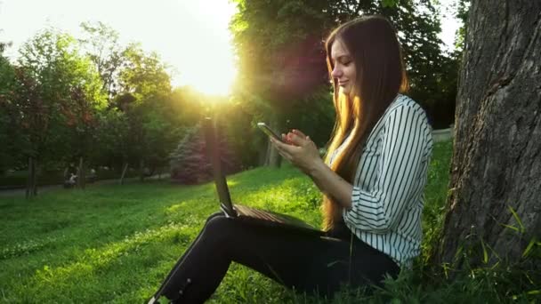 Young business woman use mobile phone while working on laptop while sitting in a park on the grass - Materiaali, video
