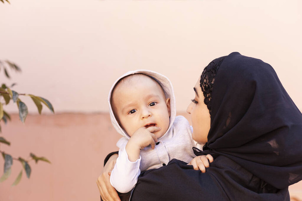 Mladá muslimka v hidžábu drží svého jednoletého chlapečka, objímá ho a líbá. Šťastná muslimská matka v abaya oblečení polibek a hrát si se svým malým dítětem - Fotografie, Obrázek