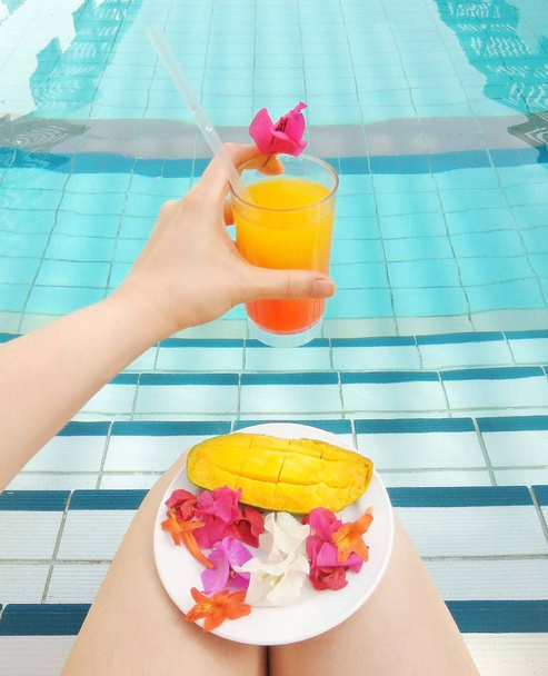 Tequila zonsopgang of sinaasappelsap in vrouwelijke hand geserveerd tropische bloem in een zwembad. Bord met bloemen bougainvillea en rijpe mango.Concept van levensstijl roeping, ontspannen, rust, spa en gezonde voeding - Foto, afbeelding