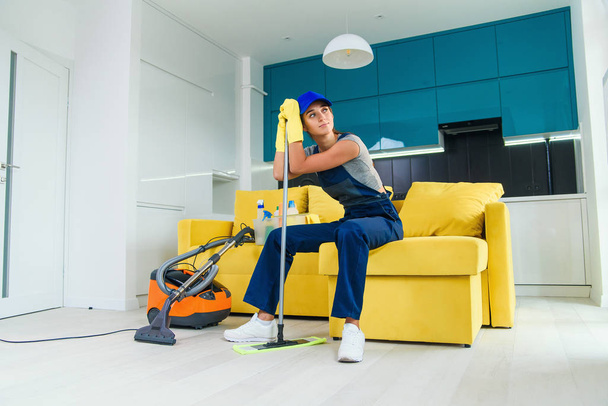 Pretty housewife tired of cleaning sits on the couch and leans to the mop after finish washing the floor. - Foto, Imagem
