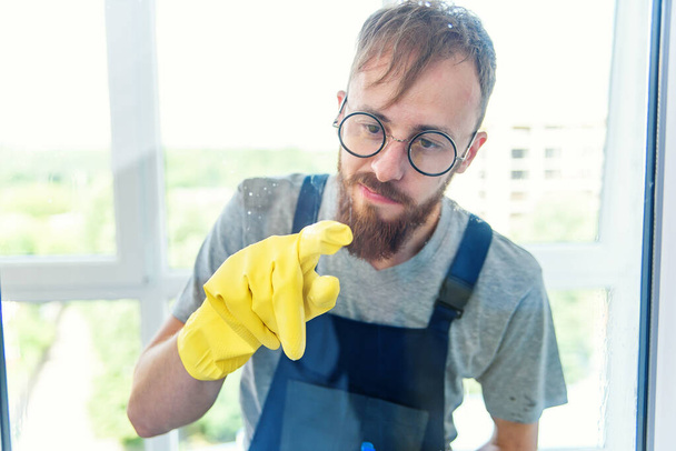 Χαρούμενος γενειοφόρος με αστεία γυαλιά καθαρίζει το παράθυρο με απορρυπαντικά και αφαιρεί τη βρωμιά με το δάχτυλό του στα γάντια. - Φωτογραφία, εικόνα