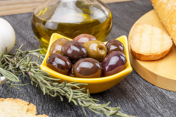 Nahaufnahme einer gelben Schüssel mit Oliven und Rosmarin. Olivenöl und Brot auf Holzgrund. - Foto, Bild
