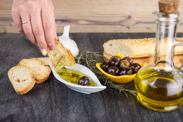 Frau taucht frisches Brot in leckeres Olivenöl mit Gewürzen in Schüssel. Olivenöl-Brotdip. Olivenöl-Sauce in weißer Schüssel & griechische Oliven auf Holz-Hintergrund.  - Foto, Bild