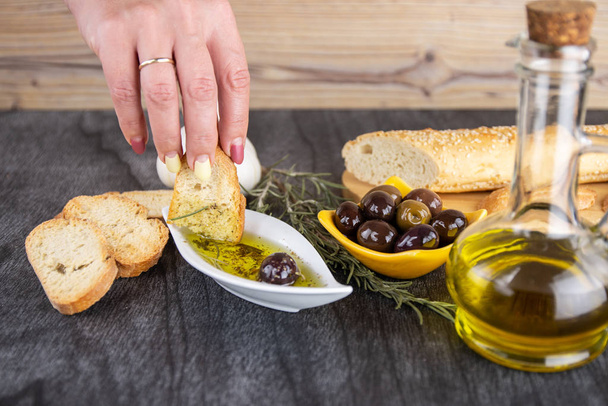 Frau taucht frisches Brot in leckeres Olivenöl mit Gewürzen in Schüssel. Olivenöl-Brotdip. Olivenöl-Sauce in weißer Schüssel & griechische Oliven auf Holz-Hintergrund.  - Foto, Bild