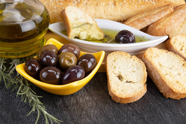 Nahaufnahme einer gelben Schüssel mit Oliven und Rosmarin. Olivenöl und Brot auf Holzboden. griechische schwarze Oliven  - Foto, Bild