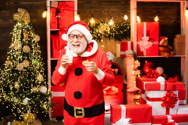Santa at home. Santa Claus enjoying in served gingerbread cake and milk. Greeting Christmas card. Happy Santa Claus eating a cookie and drinking glass of milk at home Christmas interior. - Photo, image