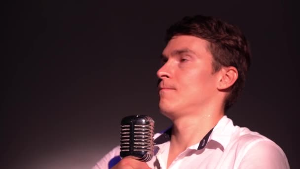 Young Caucasian man in a white open shirt goes on stage, adjusts height of classic retro glare microphone close up. Male picks up mic on scene in the dark club and says 1, 2, 3. Spotlights shine. - Footage, Video