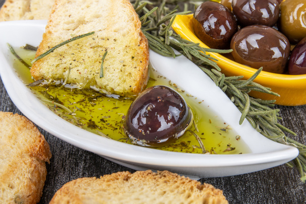 griechisches Olivenöl Brotdip. Brot mit Öl zum Dippen mit Kräutern und Gewürzen. Nahaufnahme Olivenölsosse in weißer Schüssel & griechische Oliven auf Holzgrund.  - Foto, Bild