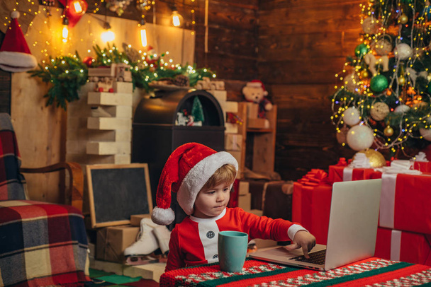 Santa helper baby boy with laptop. Bright New Years interior. Xmas tree. Indoor. Child at Christmas tree and fireplace on Xmas eve. Christmas time. Christmas attributes. - Fotografie, Obrázek
