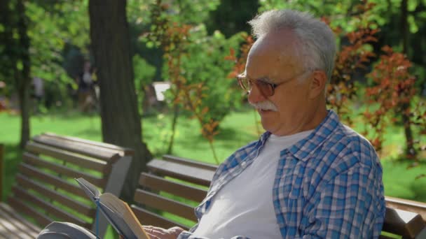 Starší muž sedící na lavičce a čtoucí v parku legrační knihu - Záběry, video