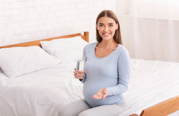 Беременная женщина с таблетками и водой сидит в спальне
 - Фото, изображение