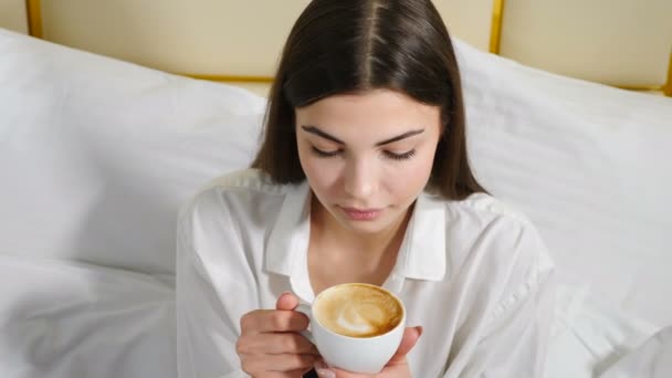 Ritratto di giovane donna attraente e sicura seduta su biancheria da letto bianca che regge la tazza con caffè e sorride guardando la macchina fotografica. Buongiorno in camera d'albergo. Girato in 4k
 - Filmati, video