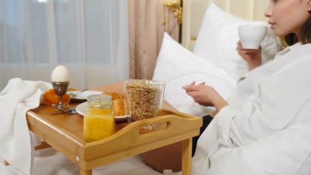 Widok z tyłu młodej kobiety ubranej w białą męską koszulę relaksującej się w łóżku z śnieżnobiałymi pościelami, jedzącej śniadanie na małej tacy. Kobieta bez twarzy delektująca się poranną kawą w świetle dnia. 4k - Materiał filmowy, wideo