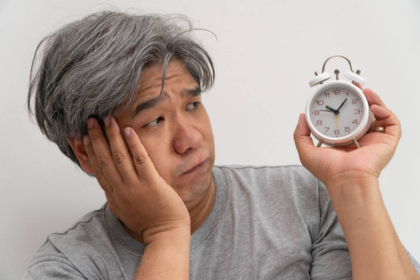 Азиатский мужчина средних лет держит белый будильник, и его лицо показало скуку и плохое самочувствие, его проблемой является расстройство сна. Концепция страдающей бессонницы
 - Фото, изображение