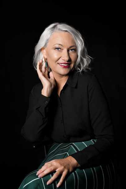 Portrait of stylish mature woman sitting on chair against dark background - Photo, image