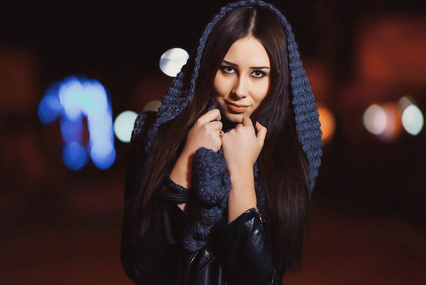 Porträt eines Mädchens mit langen schwarzen Haaren in dunkler Kleidung in einer nächtlichen Stadt. Mädchen auf dem Hintergrund von Bokeh Lichter der Nacht Stadt. eine junge Frau in schwarzer Lederjacke und Strickschal - Foto, Bild