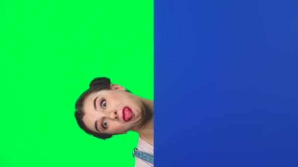 Funny girl looking out from behind blue blank placard on green screen at studio, slow motion - Felvétel, videó
