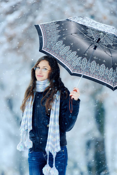 街の冬の屋外で雪の下で傘を持つ女の子。雪の下で冬の路上でのビジョンのための眼鏡のロマンチックな女の子。冬の風景の中に美しい若いブルネットの女性 - 写真・画像