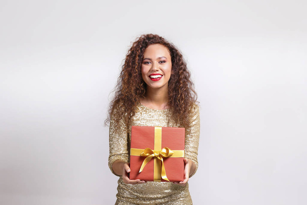 Studio portrait of young woman with dark skin and long curly hair wearing tight sexy dress, holding wrapped present with golden bow. Close up, copy space for text, isolated background. - Photo, Image