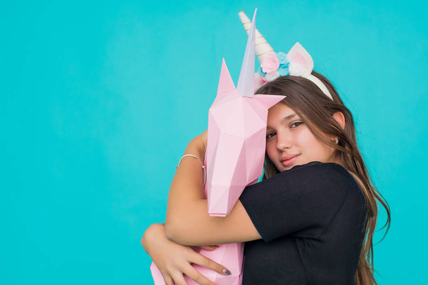 Kawaii teenage girl. Cute young woman with unicorn horn holding and hugging handmade paper pink unicorn over blue background. - 写真・画像