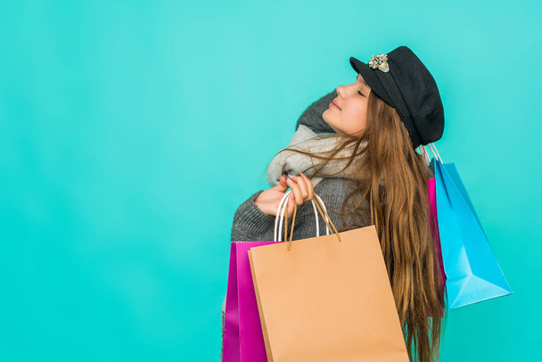 Portrait of a smiling pretty girl with shopping bags over blue background. Black friday concept. Kawaii teenage girl. Cute young woman wearing coat and wool cap. - Photo, Image