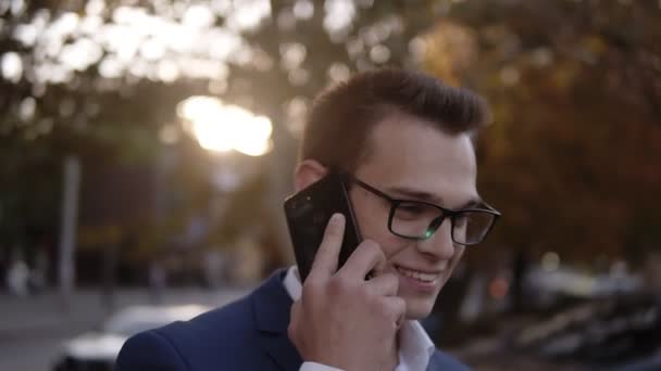 Portrait of businessman in stylish glasses talking on the mobile phone on streets of business district. Young smiling man using smart phone on the street. Sunlight and autumn trees on the background - Кадри, відео